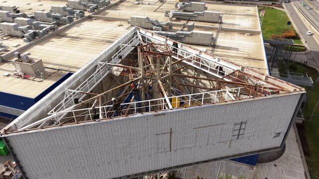 Aerial approach toward workers inside a triangular billboard structure, revealing rusted cross braces, white catwalks and a busy adjacent highway.