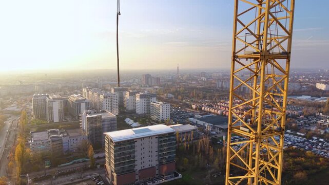 Aerial view of a large tower crane operating above a major urban construction site, with modern office buildings, residential blocks and a large sports arena visible across the surrounding cityscape.