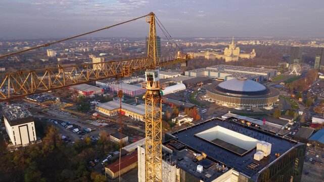Aerial drone footage of a tower crane operating above a modern high-rise construction site with a panoramic city skyline in the background. The scene showcases urban development, commercial real estat
