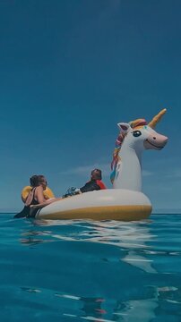 Mother son inflatable unicorn float in crystal ocean under bright blue sky, mother steadying float while child laughs, gentle waves, sunlit reflections, vibrant summer colors, vertical video