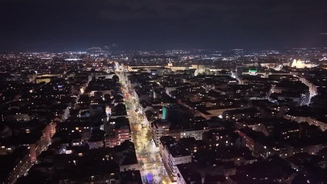 Aerial drone footage capturing New Year's Eve fireworks erupting across a brightly illuminated city at night. Multiple fireworks displays light up the urban skyline while a long boulevard stretches th