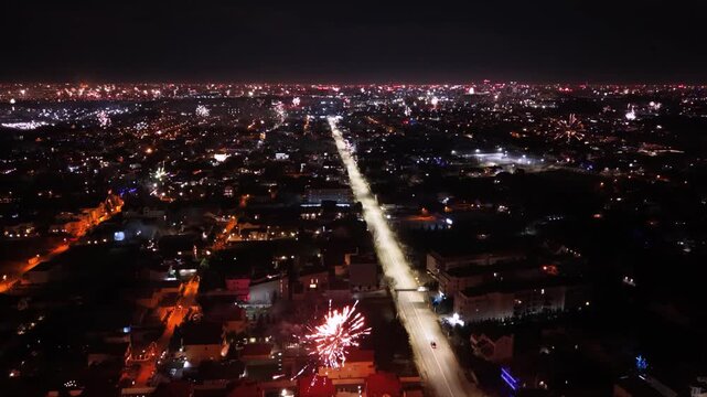 Aerial drone footage capturing New Year's Eve fireworks erupting across a brightly illuminated city at night. Multiple fireworks displays light up the urban skyline while a long boulevard stretches th