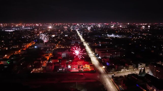 Aerial drone footage capturing New Year's Eve fireworks erupting across a brightly illuminated city at night. Multiple fireworks displays light up the urban skyline while a long boulevard stretches th