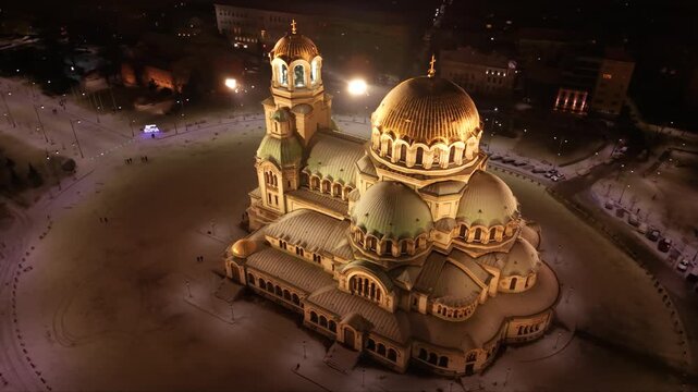 Night aerial view of a brightly lit cathedral with golden domes, surrounded by snowy grounds and city streets of Sofia, Bulgaria. Orthodox Christian church seen from above at night