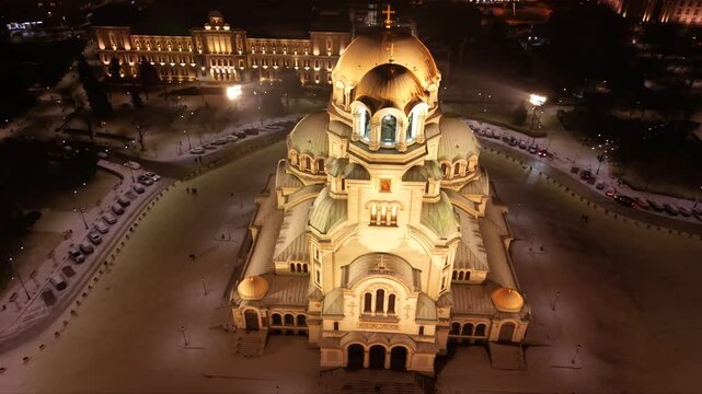 Cinematic aerial drone footage of the illuminated Alexander Nevsky Cathedral on a snowy winter night in Sofia, Bulgaria. The golden domes glow against the dark cityscape while fresh snow blankets the 