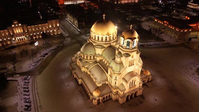 Cinematic aerial drone footage of the illuminated Alexander Nevsky Cathedral on a snowy winter night in Sofia, Bulgaria. The golden domes glow against the dark cityscape while fresh snow blankets the 