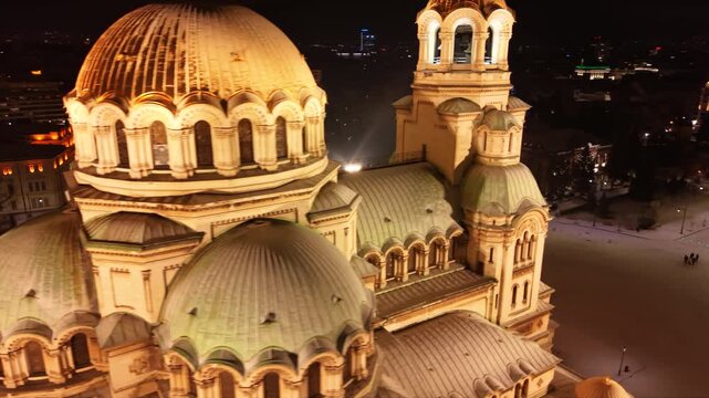 Cinematic aerial drone footage of the illuminated Alexander Nevsky Cathedral on a snowy winter night in Sofia, Bulgaria. The golden domes glow against the dark cityscape while fresh snow blankets the 