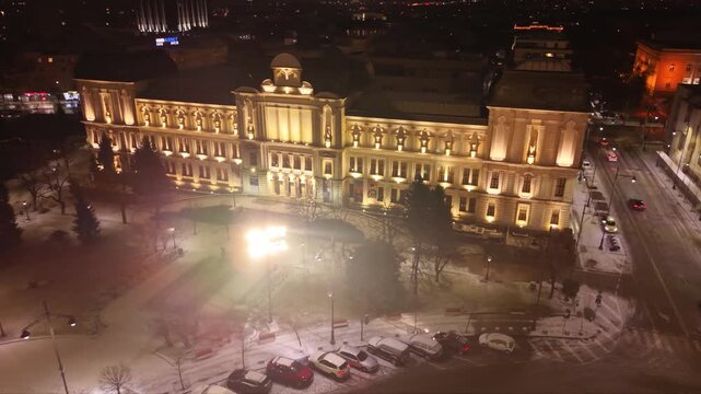 Cinematic aerial drone footage of the illuminated Alexander Nevsky Cathedral on a snowy winter night in Sofia, Bulgaria. The golden domes glow against the dark cityscape while fresh snow blankets the 