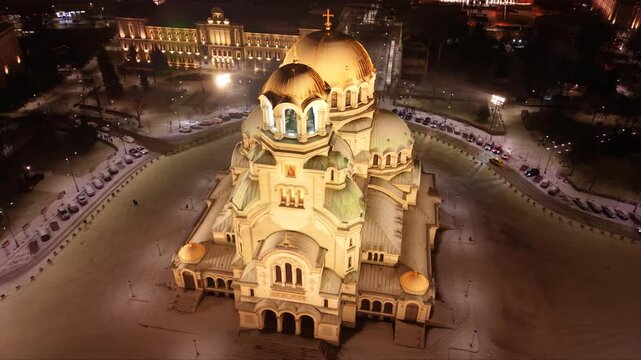 Night aerial view of a brightly lit cathedral with golden domes, surrounded by snowy grounds and city streets of Sofia, Bulgaria. Orthodox Christian church seen from above at night