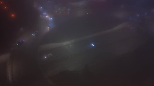 Aerial view of a fog covered urban road interchange at night, with moving headlights tracing the curved lanes around a dark central island.