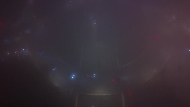 Atmospheric aerial drone footage of a city at night covered by dense fog with vehicle headlights and illuminated signs glowing through the thick mist. The low visibility creates a mysterious urban lan