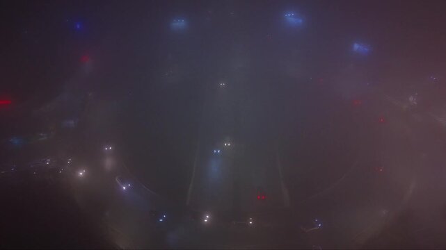 Atmospheric aerial drone footage of a city at night covered by dense fog with vehicle headlights and illuminated signs glowing through the thick mist. The low visibility creates a mysterious urban lan
