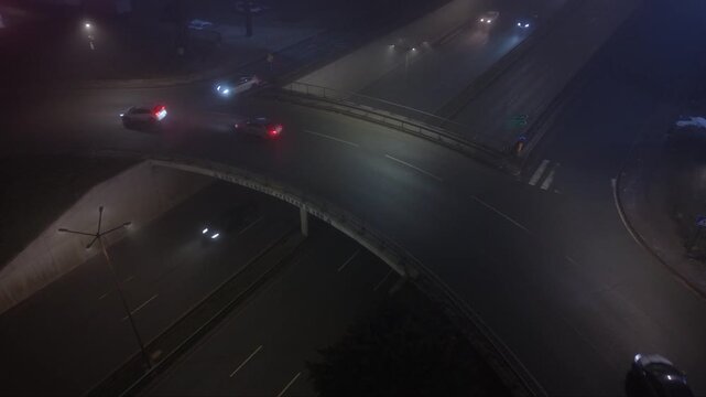 Atmospheric aerial drone footage of a city at night covered by dense fog with vehicle headlights and illuminated signs glowing through the thick mist. The low visibility creates a mysterious urban lan
