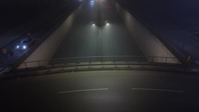 Aerial approach toward a curved bridge over a divided road at night, with cars moving on both levels beneath mist softened street lights.