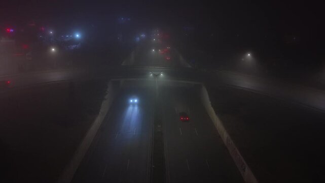 Atmospheric aerial drone footage of a city at night covered by dense fog with vehicle headlights and illuminated signs glowing through the thick mist. The low visibility creates a mysterious urban lan