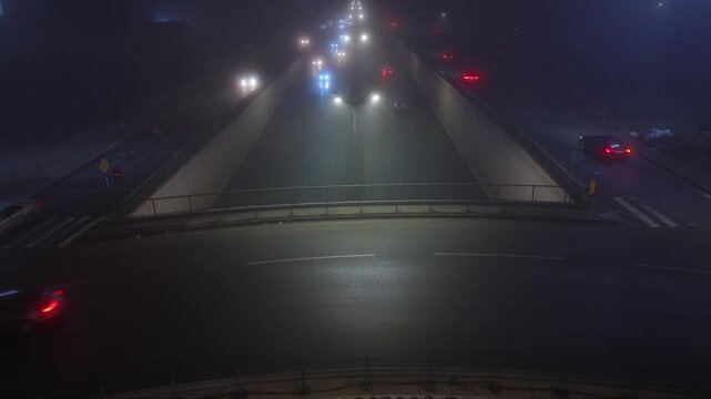 Atmospheric aerial drone footage of a city at night covered by dense fog with vehicle headlights and illuminated signs glowing through the thick mist. The low visibility creates a mysterious urban lan