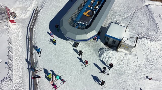 Ski lift station on a snowy slope with skiers waiting and riding, surrounded by pine trees in bright winter sunlight
