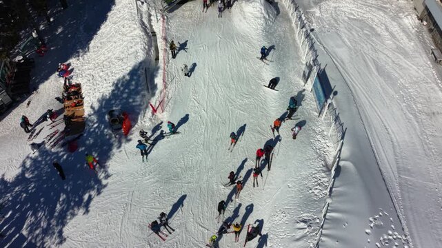 Vertical aerial drone footage of skiers and snowboarders gathering at the intersection of ski runs in a busy mountain resort. Long winter shadows stretch across freshly groomed snow as visitors prepar