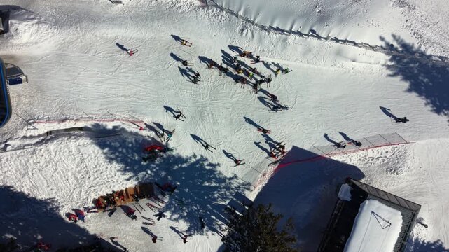 Vertical aerial drone footage of skiers and snowboarders gathering at the intersection of ski runs in a busy mountain resort. Long winter shadows stretch across freshly groomed snow as visitors prepar
