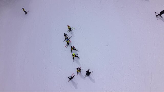 Aerial footage of a ski school lesson with beginner skiers practicing snowplow turns on a gentle slope at a mountain ski resort. The instructor leads the group downhill while other visitors enjoy wint