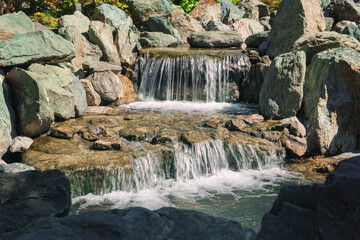 Rocky stream cascades gently over mossy stones. Sunlight glints on flowing water, soft and serene. Rugged boulders frame the natural descent. Relax, peace, calm, and quiet retreat. Horizontal photo © Andrey_Maksimov