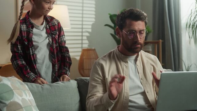 Angry Caucasian dad working at home laptop energetic girl demanding attention daughter playing distraction stressed busy father man male on couch talking noisy child kid bad behavior parenting family