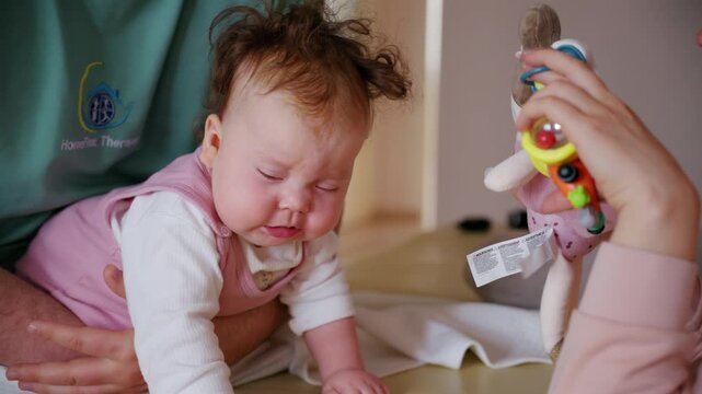A colorful baby rattle is gently presented to an infant, while caregiver is performing mobility exercises