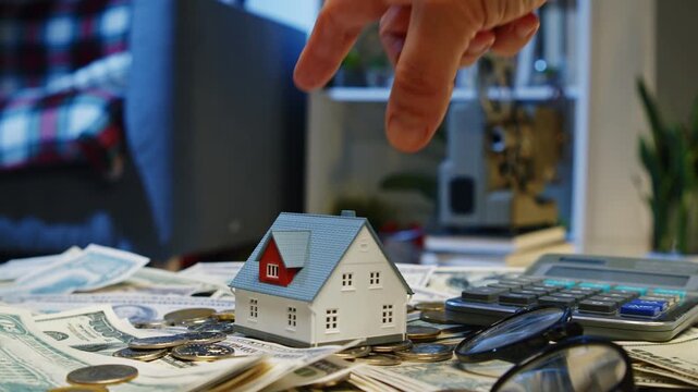 Tiny house model sits atop a pile of cash and coins in a cozy room, showcasing the connection between home ownership and financial aspirations
