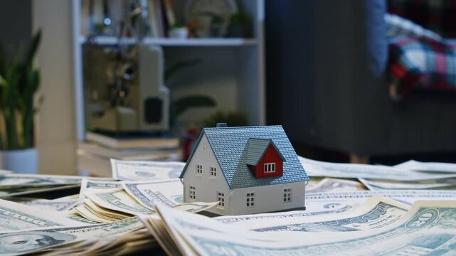A hand gently places a small house on a pile of money, symbolizing dreams of homeownership amidst a stylish interior filled with greenery and comfort