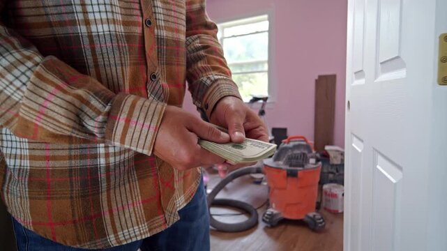 A man carefully counts dollar bills to determine how much money he needs for home repair materials. He stands in a welcoming room surrounded by tools and supplies