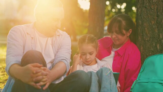 Happy family in park. Family outdoor recreation, picnic. Father mother, little daughter sitting under tree city park. People, parents, child reading book on green lawn. People relaxing. Family life