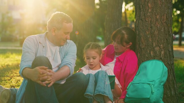 Happy family in park. Family outdoor recreation, picnic. Father mother, little daughter sitting under tree city park. People, parents, child reading book on green lawn. People relaxing. Family life