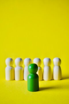 Vertical composition showing a green miniature figure standing forward out from a blurry row of white figures on a solid yellow background, signifying personal growth, advancement, and standing out.