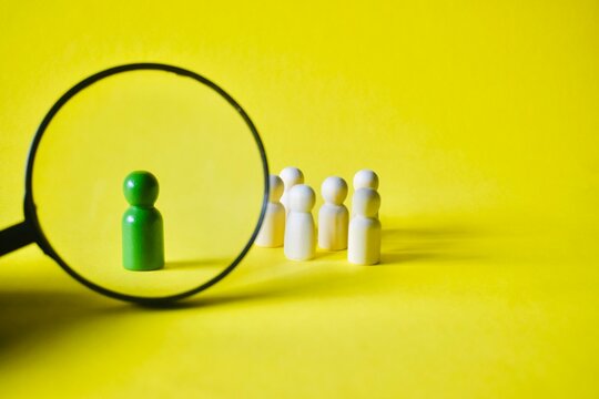 A magnifying glass zooming in on a lone green figure with an out-of-focus crowd of white figures behind it, representing executive search, unique talent scouting, and investigative focus.