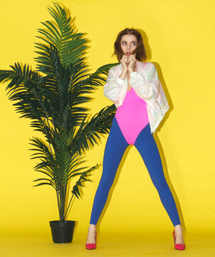 Fashion portrait of stylish young woman posing near tropical plant against bright yellow background.