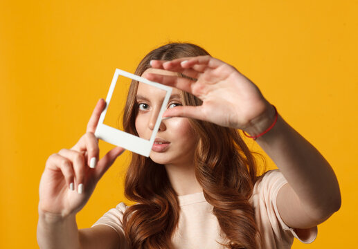 Young woman holding empty frame in front of her face against yellow background. Creative visual concept.