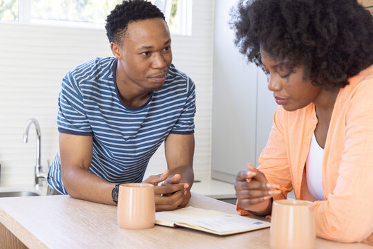 African American couple leaning over wood counter in home kitchen, reviewing notebook with pen