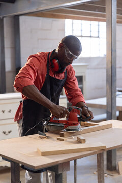 Red electric sander smoothing wooden plank on workbench in woodshop with offcuts