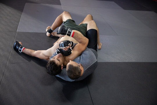 Two male grapplers grappling on padded checkered mat at dojo wearing rash guard, MMA gloves