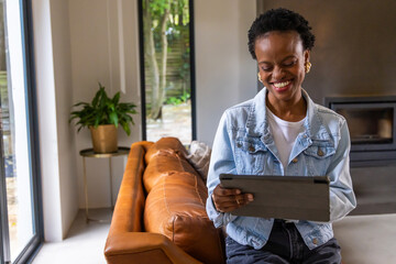 Obraz premium African American woman wearing denim jacket sitting on leather sofa holding tablet in living room