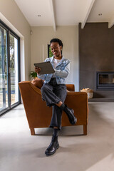 Obraz premium African American woman sitting on back of leather armchair in living room holding tablet smiling