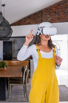 Middle-aged woman wearing white VR headset, yellow overalls, gesturing in kitchen near dining table