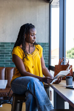 Adult African American woman in yellow blouse sitting at cafe counter reading book with coffee cup