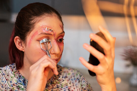 Costumed woman curling her lashes near a bloody eye wound while holding a phone