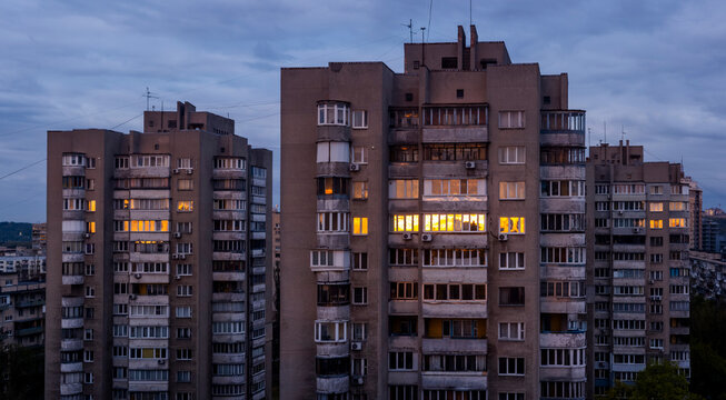 Kyiv city residential buildings standing against a cloudy evening sky, with internal lights illuminating numerous windows, symbolizing urban life and community within dense housing architecture