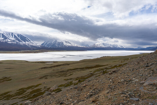 Frozen Alpine Lake with Melting Ice, Larch Forest and Snow-Capped Mountain Range Under Dramatic Clouds, Mongolian Altai