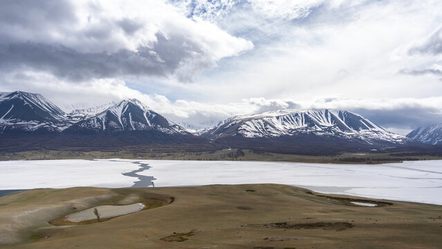 Frozen Alpine Lake with Melting Ice, Larch Forest and Snow-Capped Mountain Range Under Dramatic Clouds, Mongolian Altai