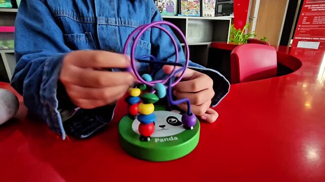 Close-up of a young child's hands wearing a denim jacket playing with a colorful wooden wire bead maze toy on a bright red table inside a play area.
