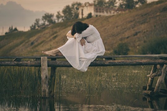 Authentic barefooted young woman in vintage white dress sits curled up on old wooden small bridge over a calm water in summer. Atmosphere of pagan witchcraft, earth magic and medieval