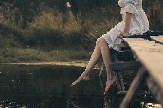 Atmospheric scene of faceless young woman in white vintage dress sitting and swinging her bare legs on old wooden pier over dark wild pond in summer